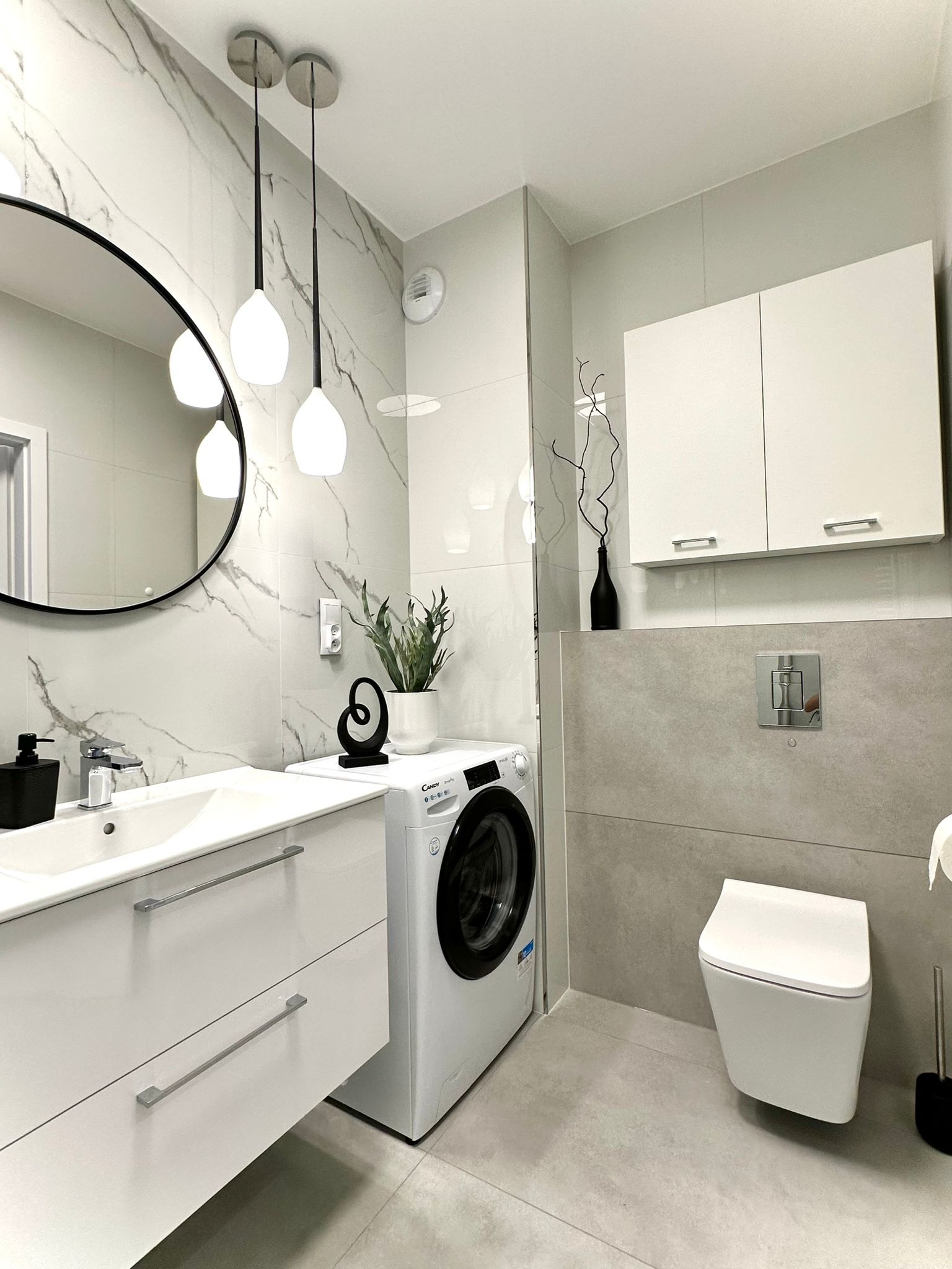 Nowoczesna łazienka w odcieniach szarości i bieli z owalnym lustrem, wiszącą toaletą, pralką Candy i dekoracjami.