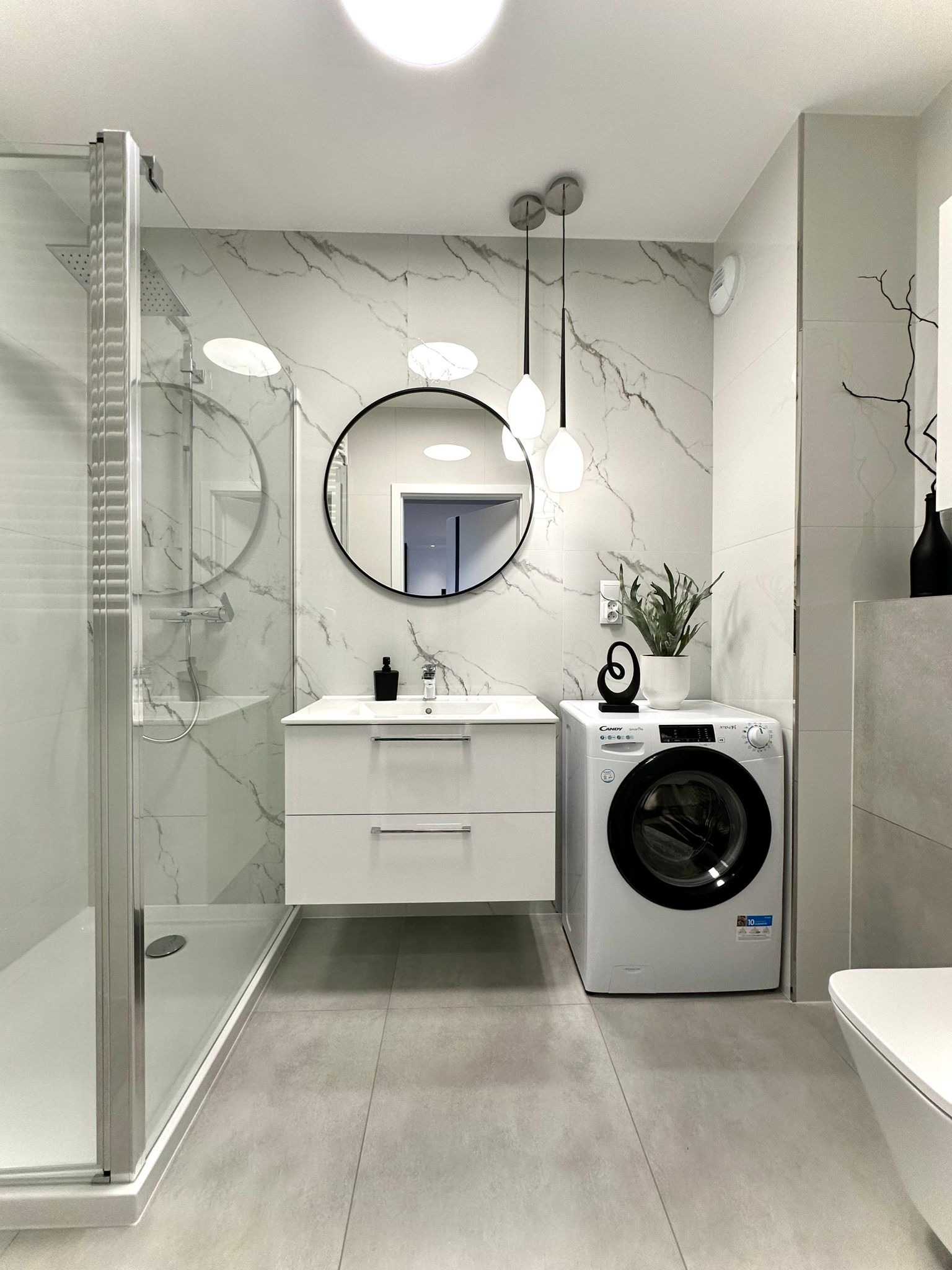 Nowoczesna łazienka z kabiną prysznicową, umywalką z szafką, pralką Candy i okrągłym lustrem, wykończona płytkami imitującymi marmur.