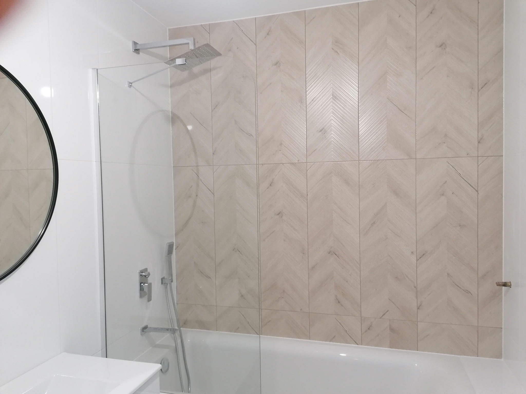 Nowoczesna łazienka z beżowymi płytkami ułożonymi w jodełkę, szklana kabina prysznicowa, bateria podtynkowa i okrągłe lustro z czarną ramą.