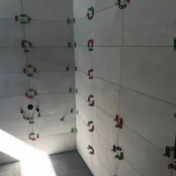 Świeżo ułożone, szare płytki w łazience, utrzymywane w równych odstępach przy pomocy kolorowych klipsów i klinów poziomujących, widoczne czarne sznurki wyznaczające spoiny.