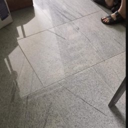 U jacka - Jasnoszara, polerowana podłoga wyłożona kwadratowymi płytkami z widocznym rysunkiem kamienia, fragment drewnianej skrzynki na narzędzia i stopy w sandałach w tle.