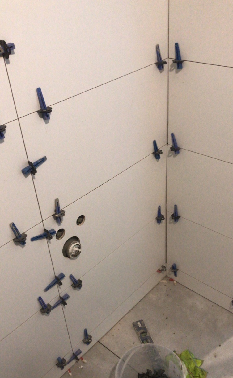 Układanie jasnych płytek ściennych w narożniku łazienki, z użyciem niebieskich klipsów poziomujących i krzyżyków dystansowych, widoczny otwór na instalację sanitarną oraz wiadro z zapasem klipsów.