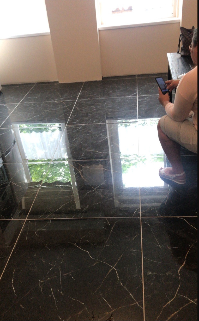 Błyszcząca, czarna podłoga z marmurowym wzorem i jasnymi fugami, odbijająca okno z widokiem na zieleń; osoba siedząca na brzegu, przeglądająca telefon.