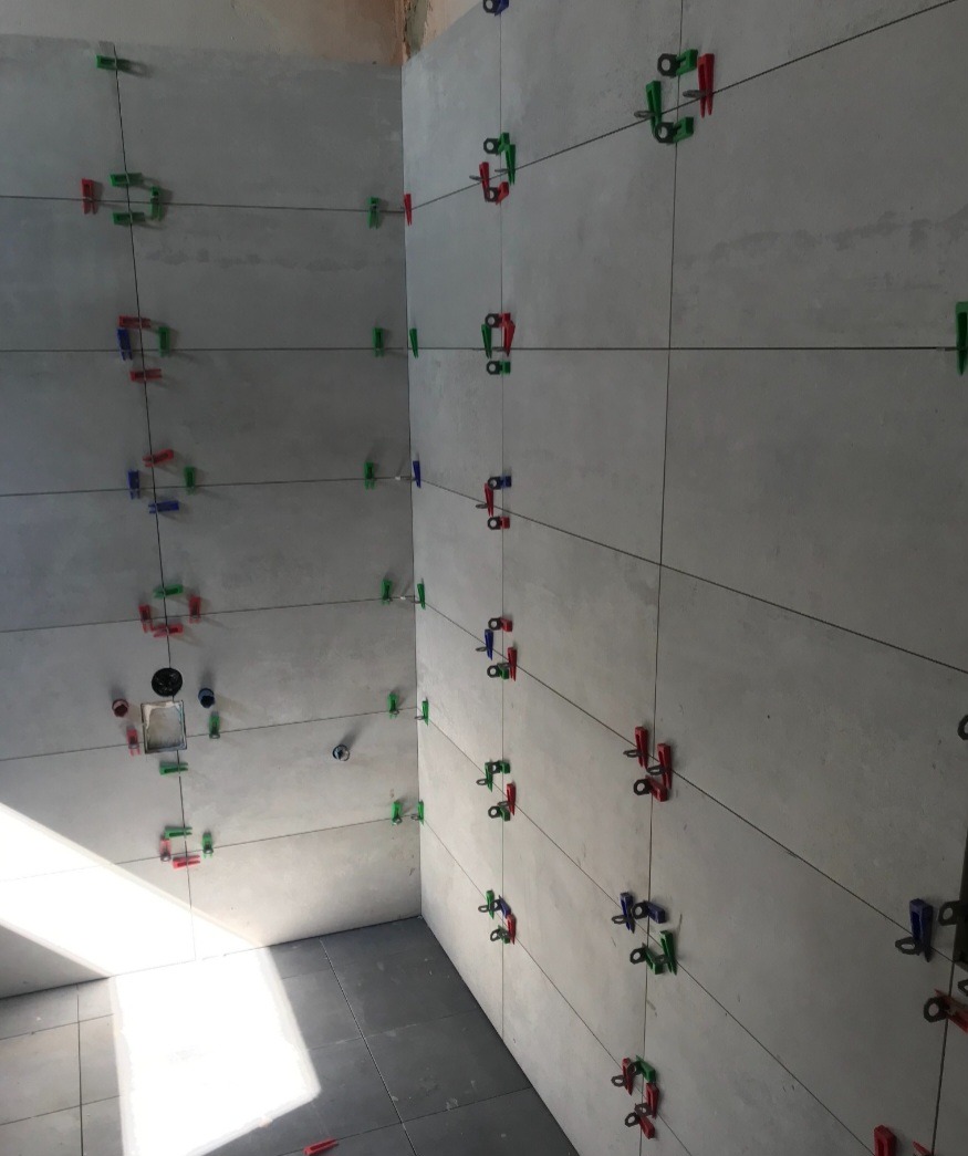 Świeżo ułożone, szare płytki w łazience, utrzymywane w równych odstępach przy pomocy kolorowych klipsów i klinów poziomujących, widoczne czarne sznurki wyznaczające spoiny.