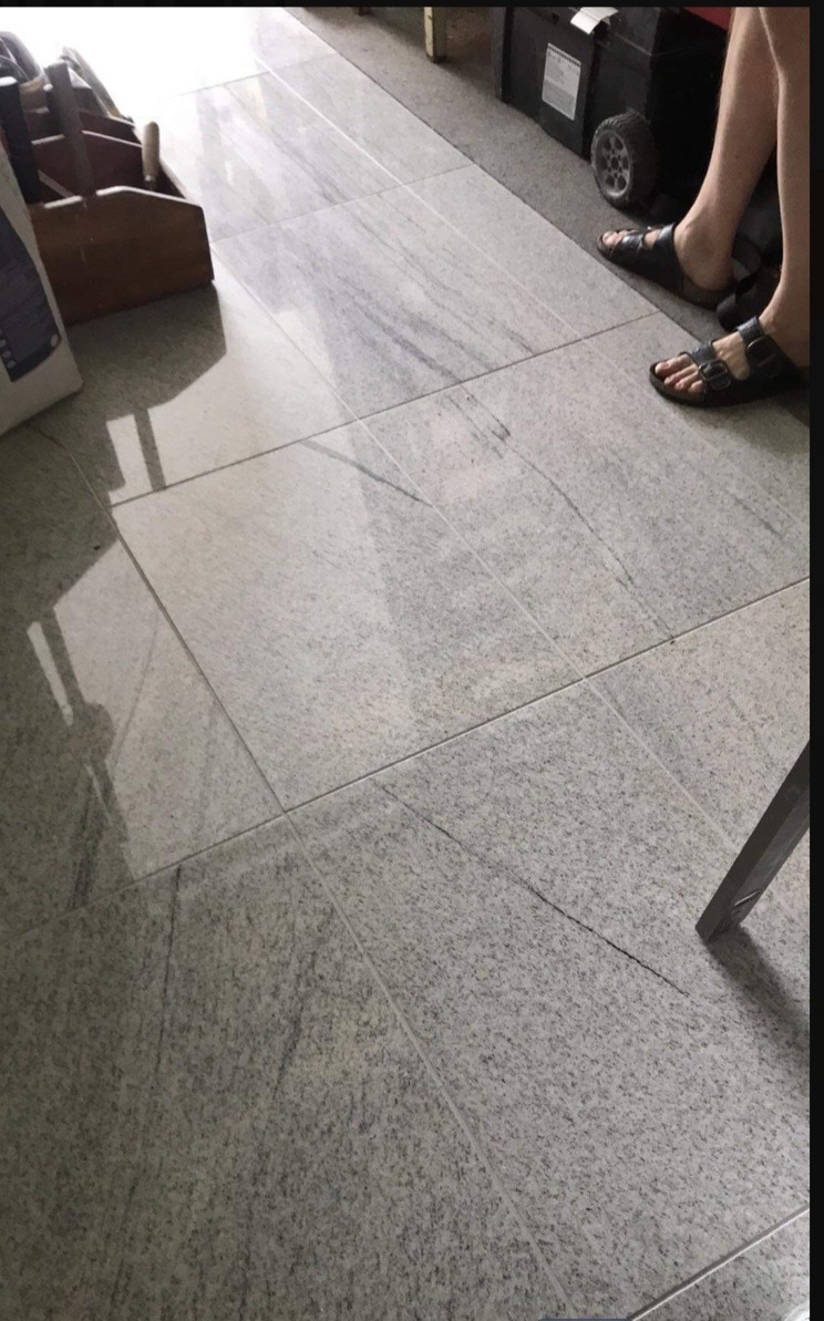 Jasnoszara, polerowana podłoga wyłożona kwadratowymi płytkami z widocznym rysunkiem kamienia, fragment drewnianej skrzynki na narzędzia i stopy w sandałach w tle.