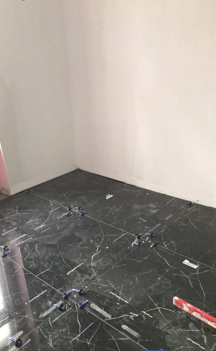 Układanie czarnych płytek imitujących marmur na podłodze w rogu pomieszczenia, z użyciem niebieskich krzyżyków dystansowych i poziomicy.