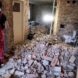 JURASSIC BUD YURII PUKHOV - Wnętrze w trakcie demontażu: sterta gruzu ceglanego na podłodze, odsłonięte cegły na ścianach, widoczna instalacja sanitarna, oświetlenie robocze i fragment sylwetki osoby w czerwonym kombinezonie.