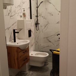 JURASSIC BUD YURII PUKHOV - Nowoczesna łazienka w trakcie wykańczania: białe marmurowe płytki, drewniana szafka pod umywalką z czarną armaturą, czarna deszczownica. Widoczny sprzęt budowlany.