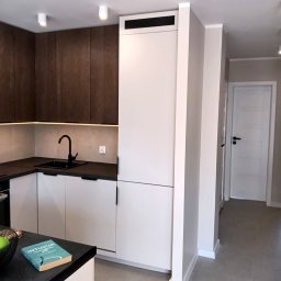 JURASSIC BUD YURII PUKHOV - Nowoczesna kuchnia z białymi szafkami i ciemnym blatem, oświetlenie LED pod szafkami wiszącymi. Minimalistyczny design z czarną armaturą i książką na blacie.