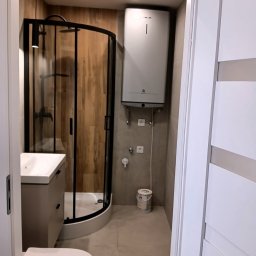 JURASSIC BUD YURII PUKHOV - Nowoczesna łazienka po remoncie: kabina prysznicowa z czarną ramą, umywalka z szafką w odcieniach szarości, biała toaleta i bojler. Widok zza otwartych, białych drzwi.