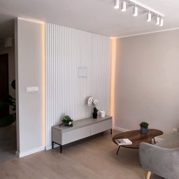 JURASSIC BUD YURII PUKHOV - Nowoczesny salon z lamelami ściennymi podświetlonymi LED, szarą komodą i drewnianym stolikiem kawowym. Minimalistyczny design, jasne kolory, oświetlenie punktowe.