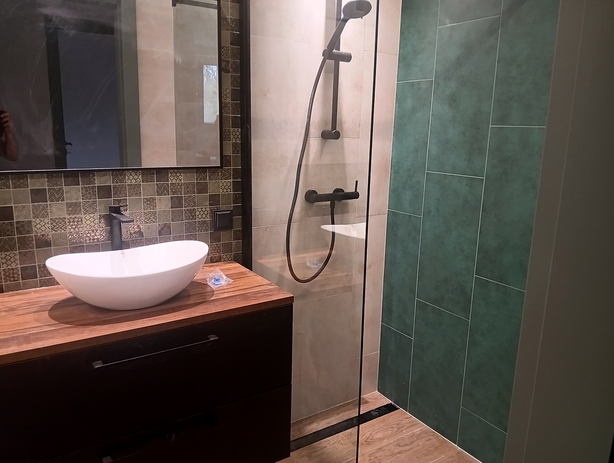 Nowoczesna łazienka z owalną umywalką nablatową, drewnianym blatem, mozaiką na ścianie, kabiną prysznicową typu walk-in i zielonymi płytkami.