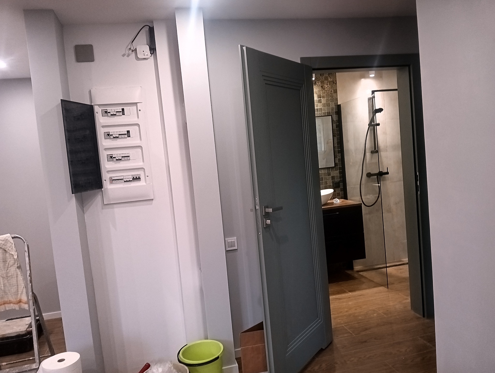 Otwarta szafka elektryczna na ścianie, drabina malarska, otwarte drzwi do łazienki z kabiną prysznicową i umywalką, widok na remontowane wnętrze.