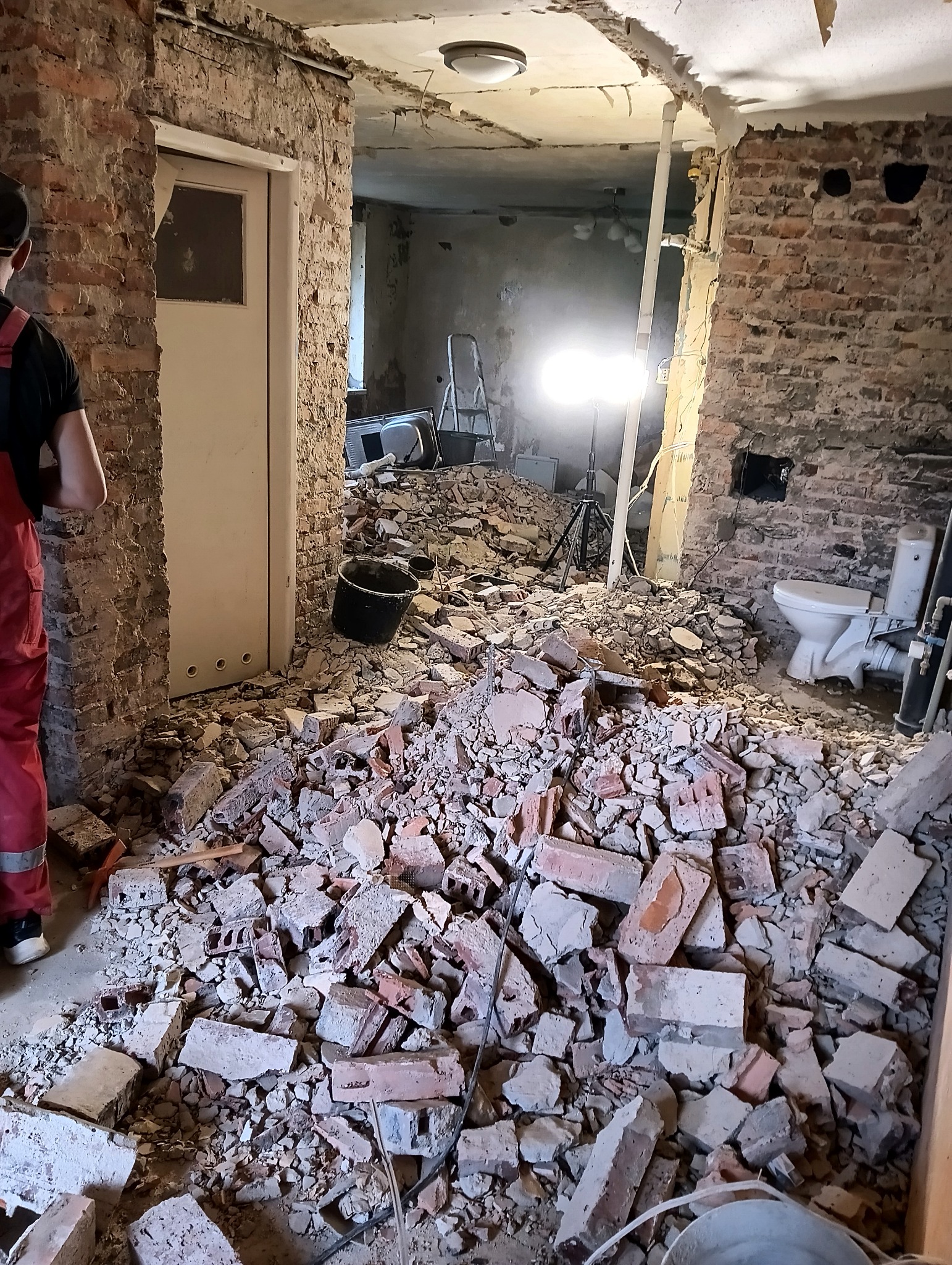 Wnętrze w trakcie demontażu: sterta gruzu ceglanego na podłodze, odsłonięte cegły na ścianach, widoczna instalacja sanitarna, oświetlenie robocze i fragment sylwetki osoby w czerwonym kombinezonie.
