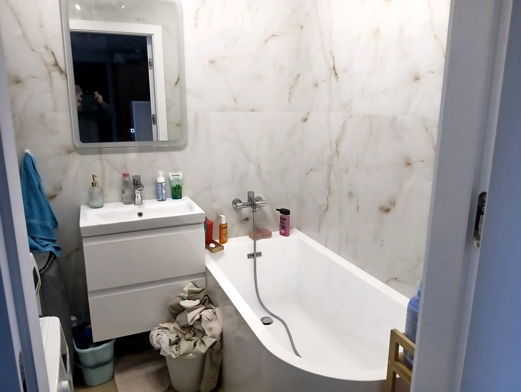 Biała łazienka z wanną i umywalką na szafce, wykończona płytkami imitującymi marmur, z widocznym odbiciem osoby robiącej zdjęcie w lustrze.