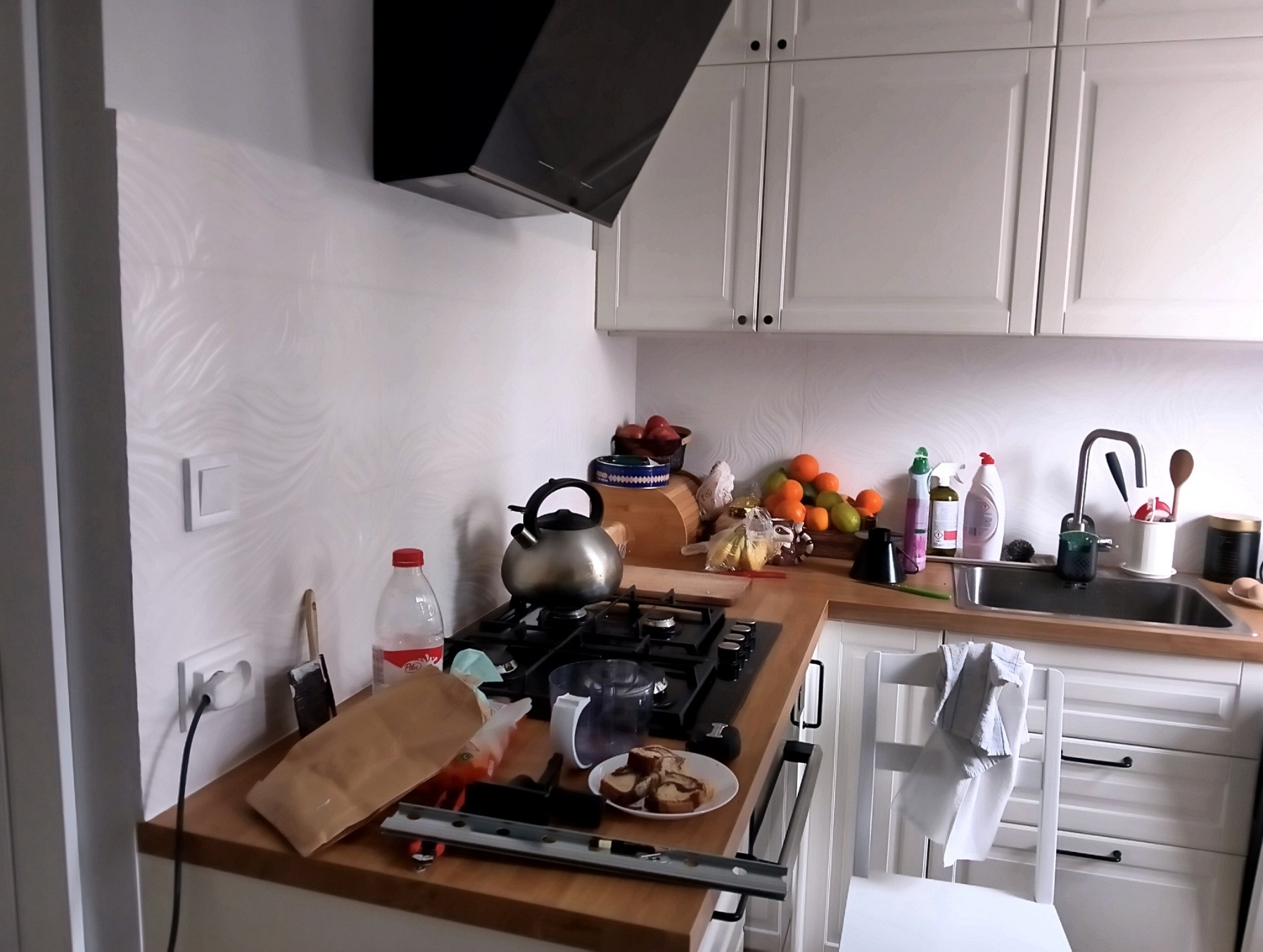 Kuchnia z białymi szafkami, czarnym okapem i blatem z drewna. Na blacie stoi czajnik, owoce, naczynia i akcesoria kuchenne. Widoczny bałagan na blacie i krzesło. Na ścianie widoczna tapeta ze wzorem.