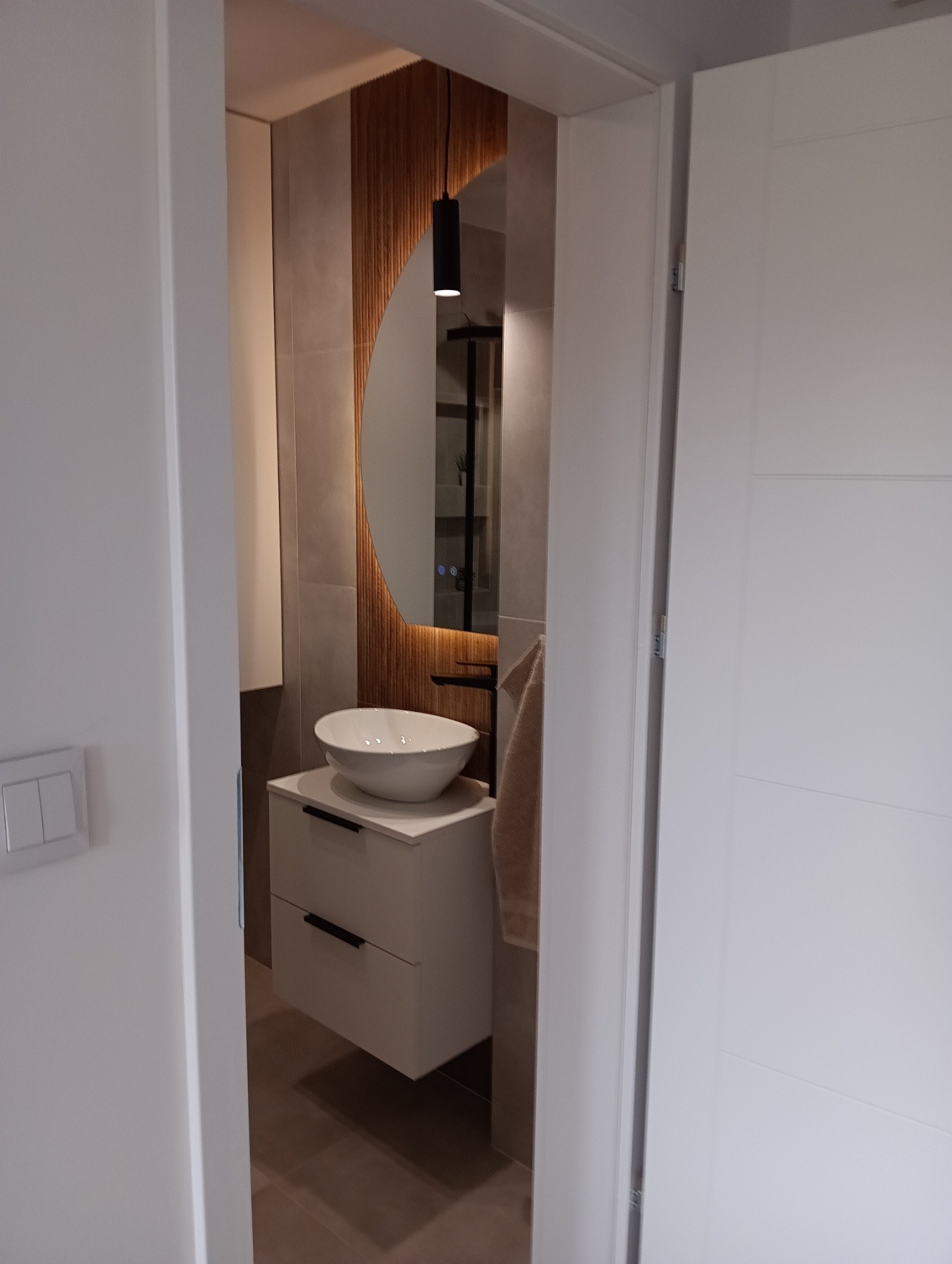 Nowoczesna łazienka z okrągłym lustrem i białą umywalką nablatową na szafce z szufladami, widziana zza uchylonych drzwi. Minimalistyczny design, ciepłe oświetlenie.