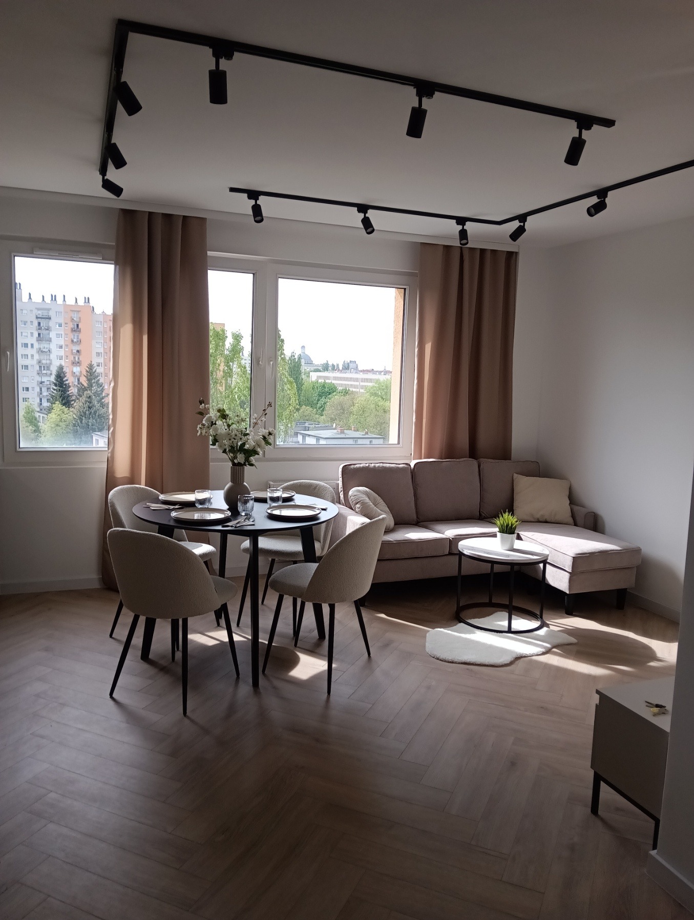 Jasne wnętrze: parkiet w jodełkę, stół z kwiatami, sofa z poduszką. Okna z widokiem na miasto. Czarny system oświetlenia szynowego pod sufitem.