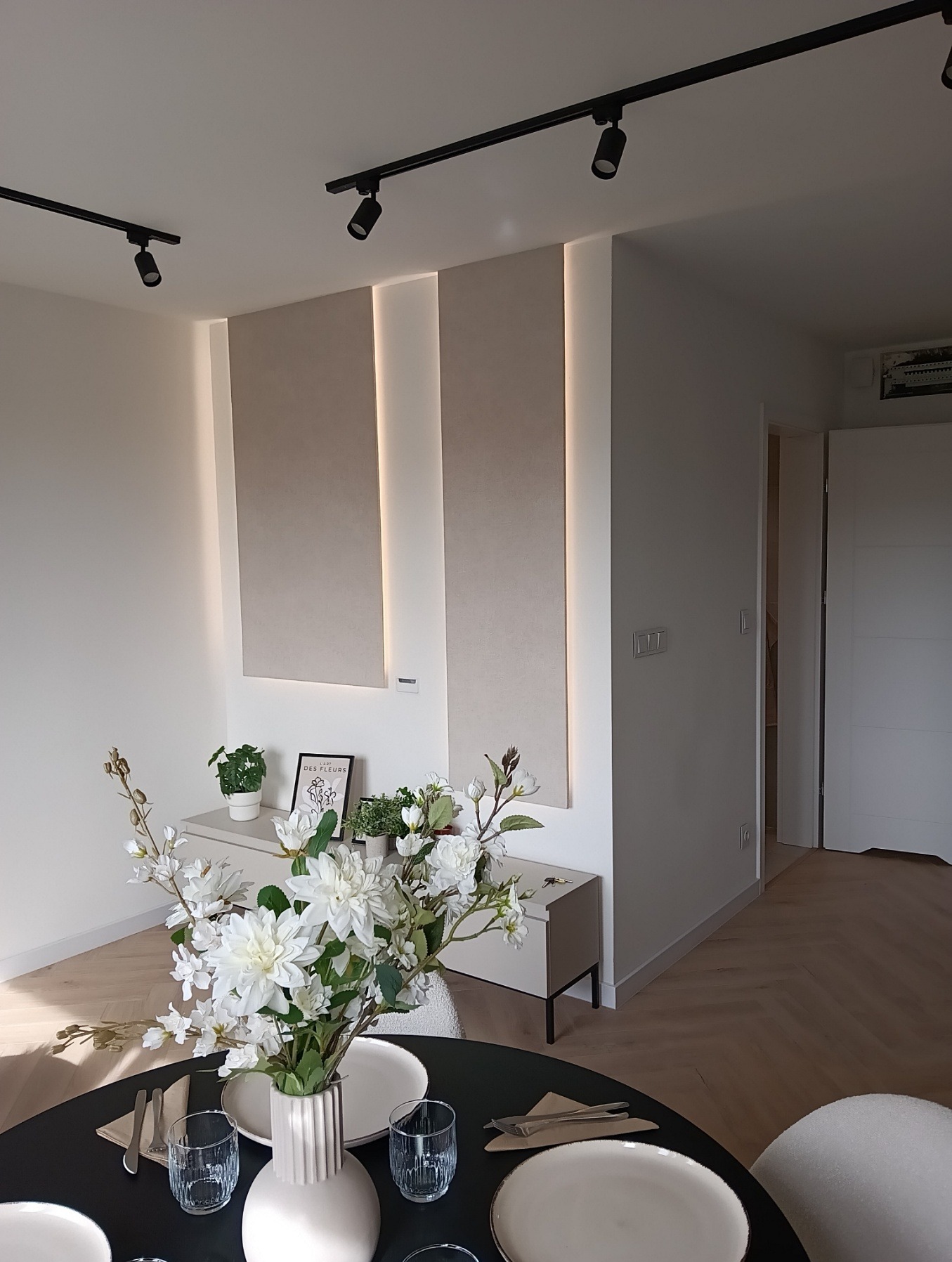 Eleganckie wykończenie wnętrza: panele ścienne z podświetleniem LED w beżowym odcieniu, czarny stół z bukietem białych kwiatów, parkiet ułożony w jodełkę.