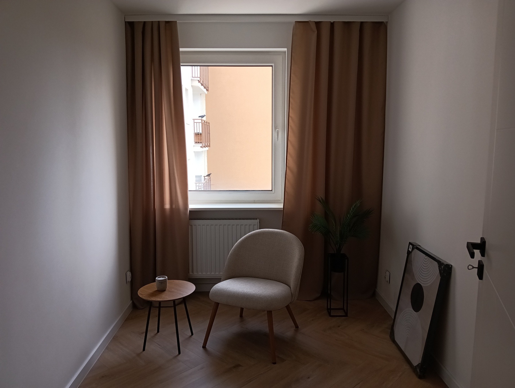 Jasne wnętrze pokoju z drewnianą podłogą, beżowym fotelem i zasłonami w oknie. Na małym stoliku stoi świeczka, a obok drzwi oparty jest obraz.