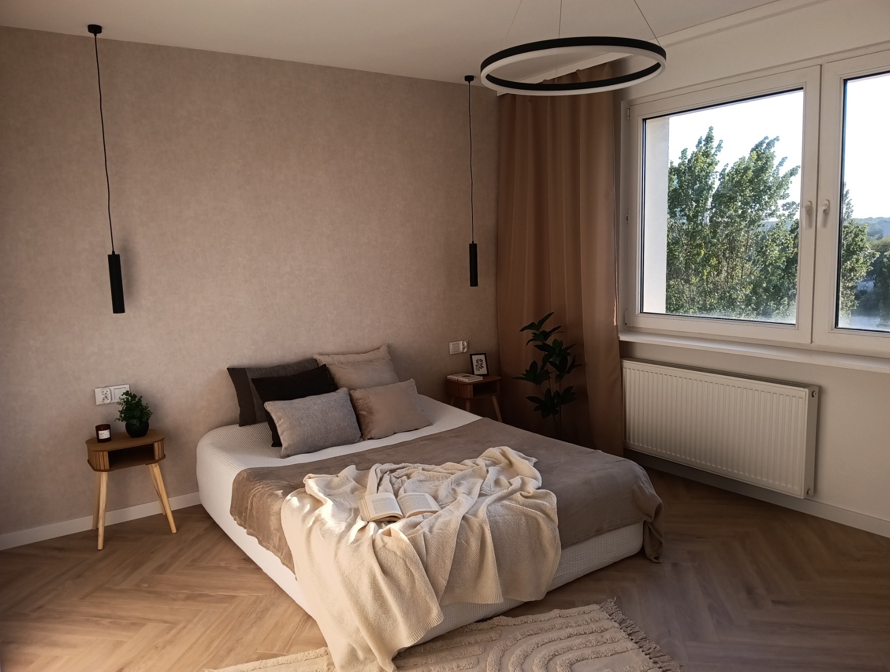 Beżowa sypialnia z parkietem w jodełkę. Na łóżku leży otwarta książka i miękki koc. Okno z widokiem na zieleń. Minimalistyczne lampy wiszące i okrągły żyrandol.