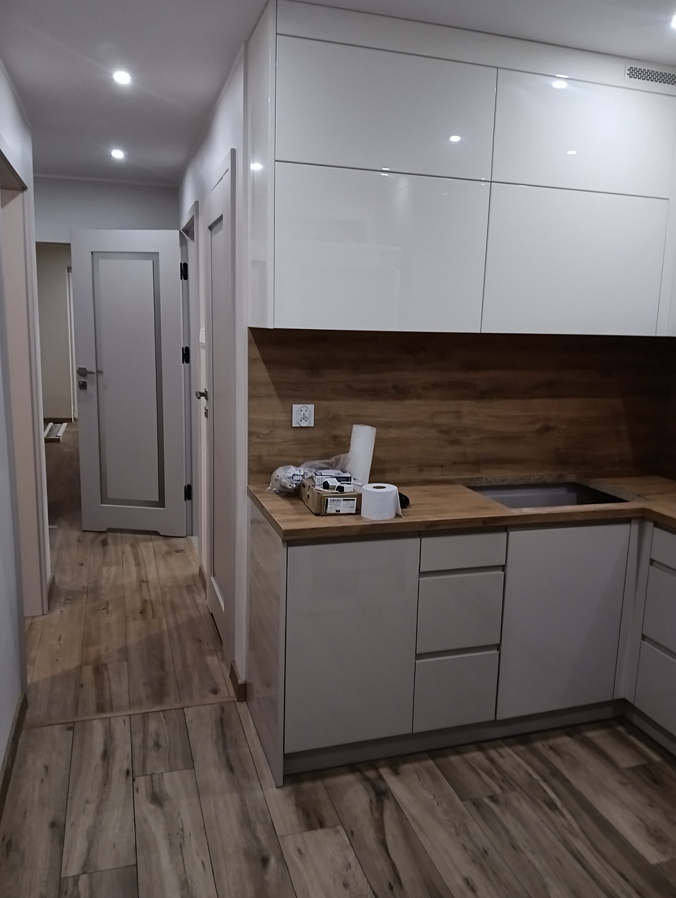 Nowoczesna kuchnia z białymi, lakierowanymi szafkami i drewnianym blatem. Widoczne akcesoria kuchenne i drewniana podłoga. W tle drzwi do innego pomieszczenia.