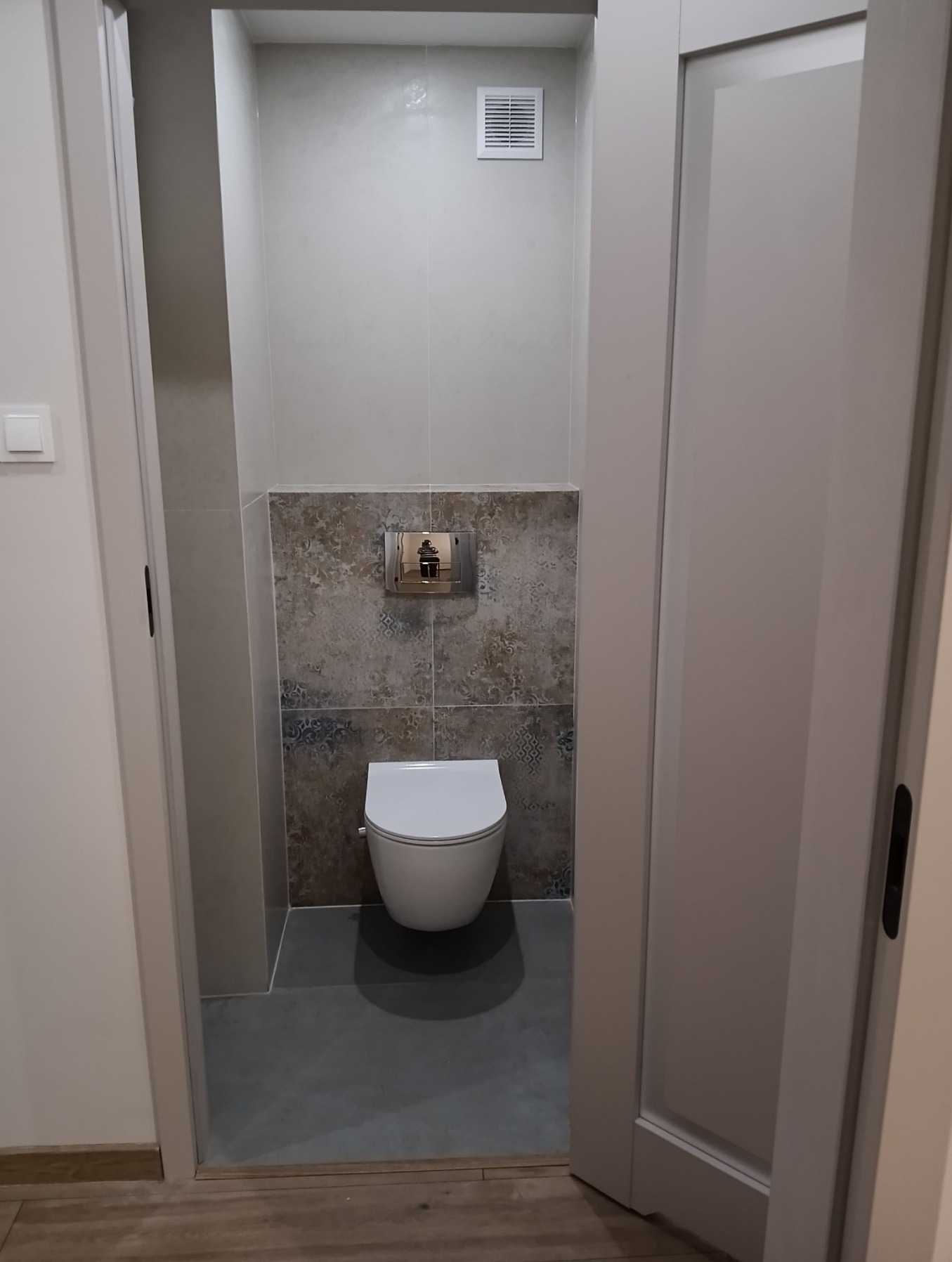 Wykończona łazienka z wiszącą toaletą, szarą podłogą i dekoracyjnymi płytkami za spłuczką. Widok zza otwartych drzwi przesuwnych.