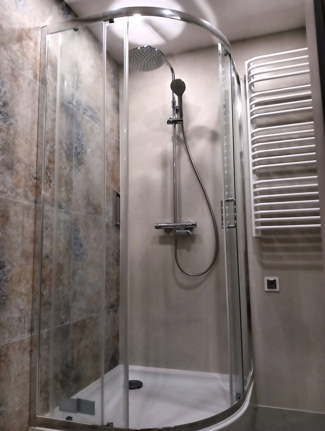 Nowoczesna kabina prysznicowa z deszczownicą i grzejnikiem drabinkowym w wykończonej łazience. Płytki imitujące kamień na ścianie.