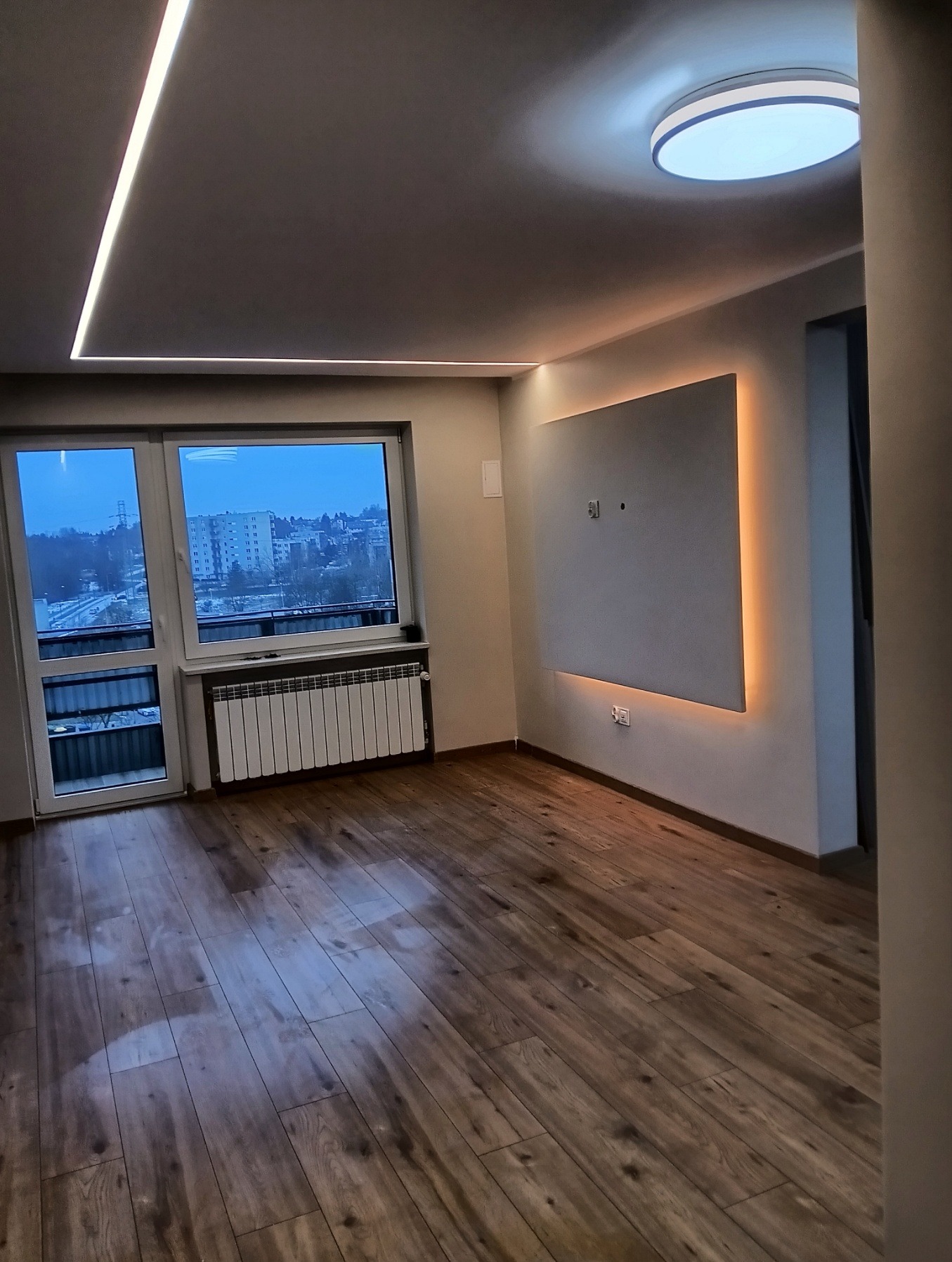 Nowoczesne wykończenie wnętrza: drewniana podłoga, podświetlany panel ścienny i sufit z oświetleniem LED. Widok na miasto przez okno.