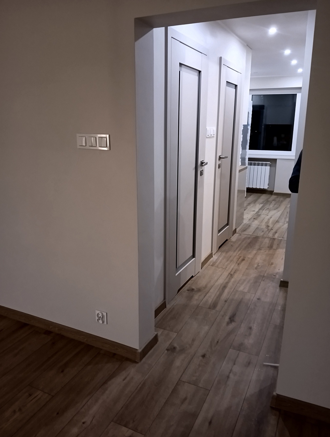 Wykończony korytarz z drewnopodobną podłogą, białymi drzwiami z szarymi wstawkami i włącznikami światła na ścianie. Widok na dalszą część mieszkania.
