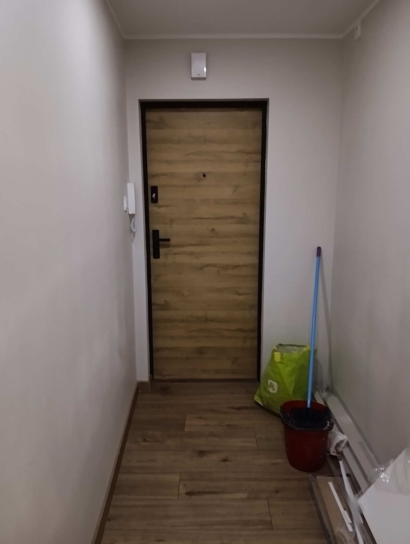 Jasne wnętrze po remoncie: drewnopodobna podłoga, drzwi z okleiną w odcieniach brązu, świeżo pomalowane ściany. Widoczne akcesoria do sprzątania.