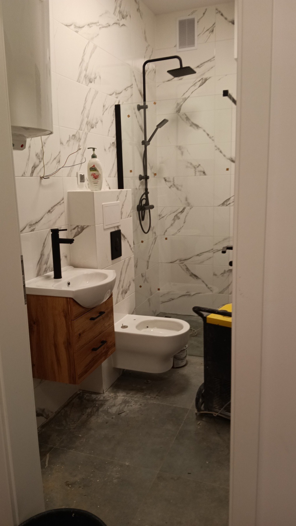 Nowoczesna łazienka w trakcie wykańczania: białe marmurowe płytki, drewniana szafka pod umywalką z czarną armaturą, czarna deszczownica. Widoczny sprzęt budowlany.