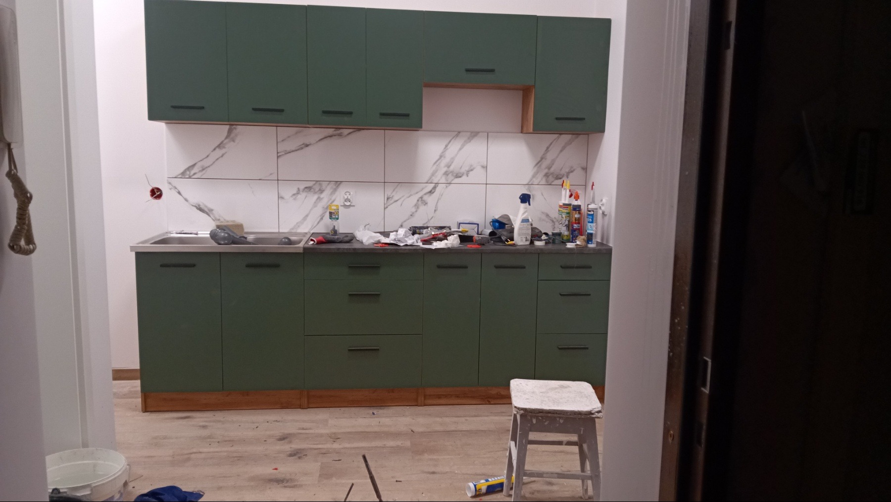 Montaż zielonej zabudowy kuchennej z marmurowym backsplashiem. Widoczne narzędzia i akcesoria montażowe na blacie. W tle fragment otwartych drzwi.