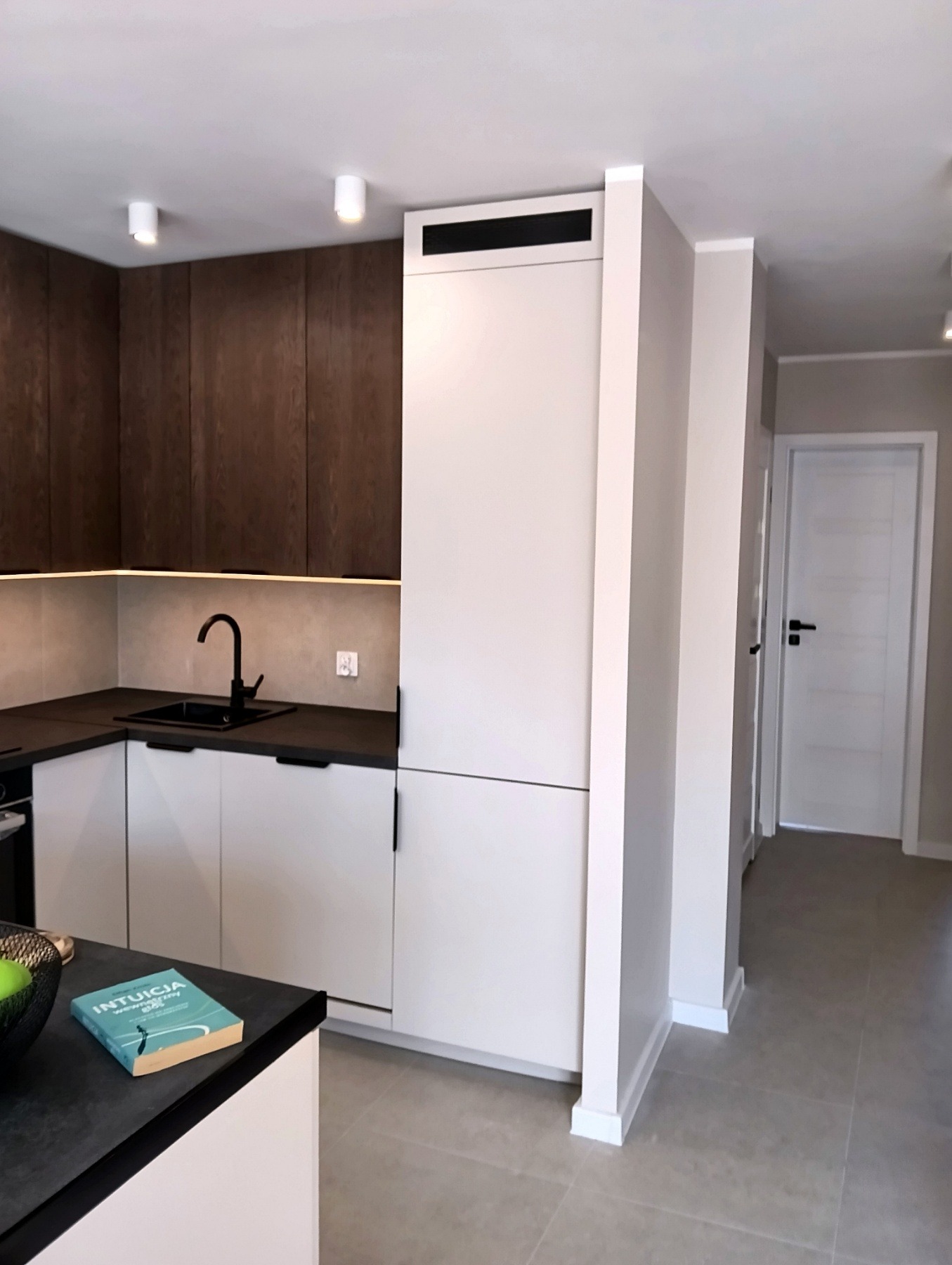 Nowoczesna kuchnia z białymi szafkami i ciemnym blatem, oświetlenie LED pod szafkami wiszącymi. Minimalistyczny design z czarną armaturą i książką na blacie.