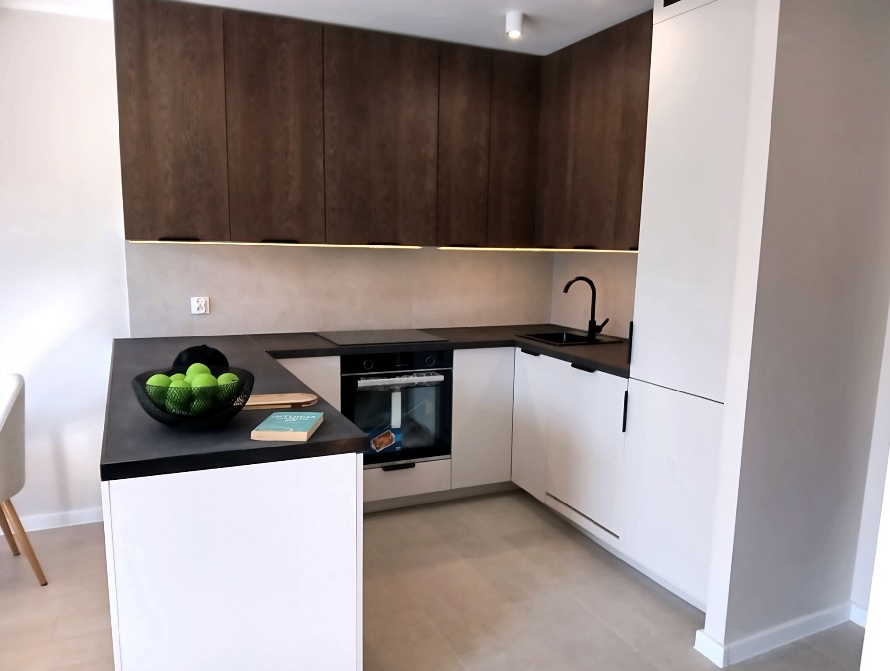 Nowoczesna kuchnia w bieli i brązie z czarnymi akcentami. Minimalistyczny design, oświetlenie LED pod szafkami, owoce w misie na blacie. Ujęcie z perspektywy.