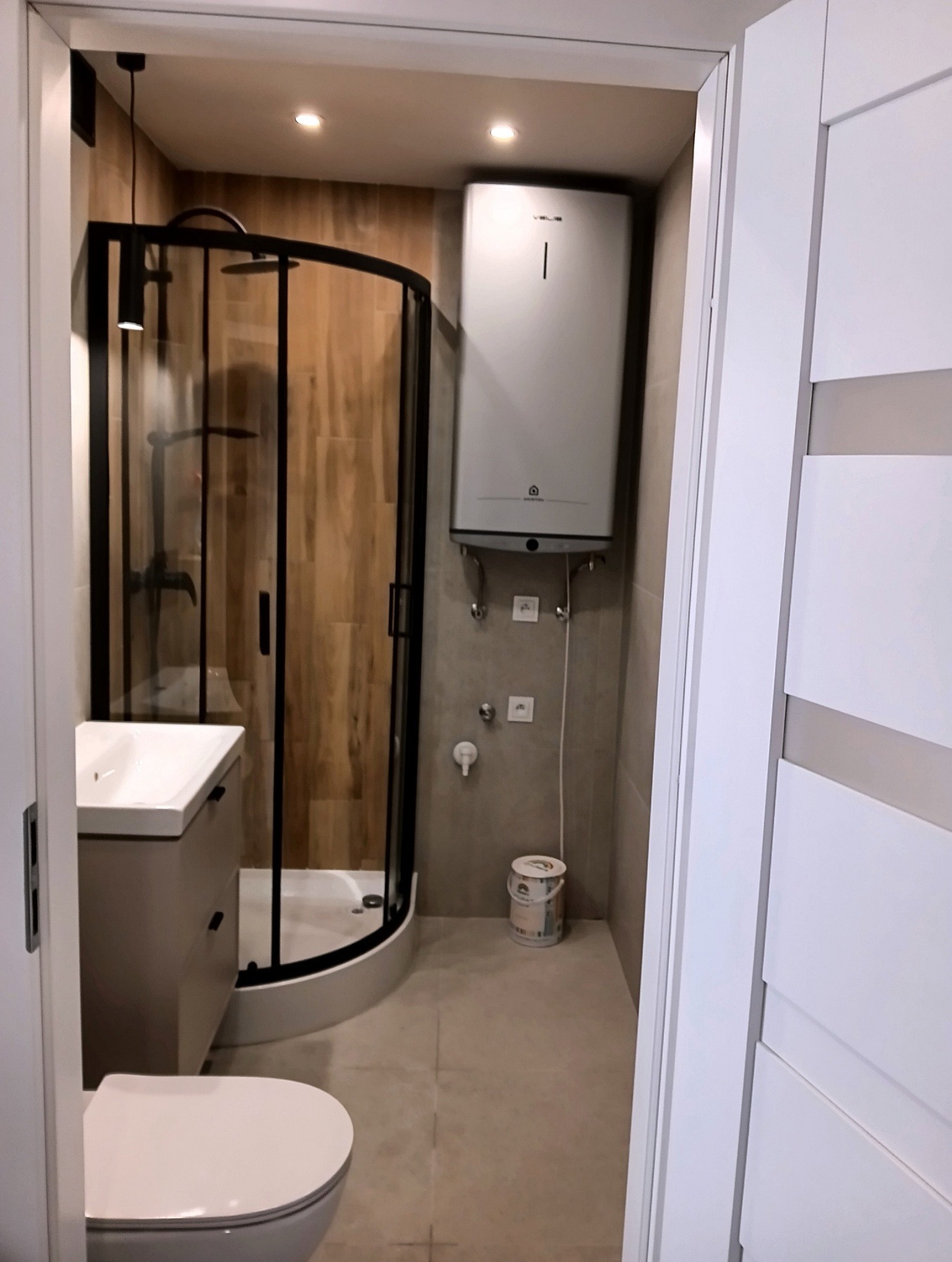 Nowoczesna łazienka po remoncie: kabina prysznicowa z czarną ramą, umywalka z szafką w odcieniach szarości, biała toaleta i bojler. Widok zza otwartych, białych drzwi.