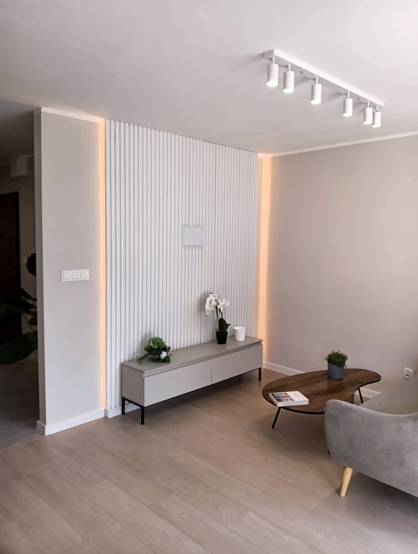 Nowoczesny salon z lamelami ściennymi podświetlonymi LED, szarą komodą i drewnianym stolikiem kawowym. Minimalistyczny design, jasne kolory, oświetlenie punktowe.