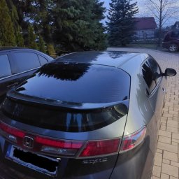 Salon auto wrappingu - Szary samochód marki Honda Civic z przyciemnionymi szybami, zaparkowany na kostce brukowej przed domem i drzewami. Widoczne tylne światła, logo Hondy i tablica rejestracyjna (częściowo zamazana).
