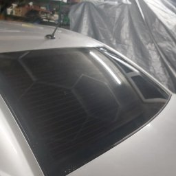 Salon auto wrappingu - Przyciemniona tylna szyba białego samochodu, widoczne delikatne zarysowania na lakierze i tło garażowe z przykrytym plandeką obiektem.