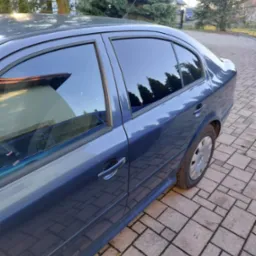 Niebieski samochód osobowy z przyciemnionymi szybami, zaparkowany na brukowanym podjeździe, widok z boku, z odbiciem drzew w szybach.