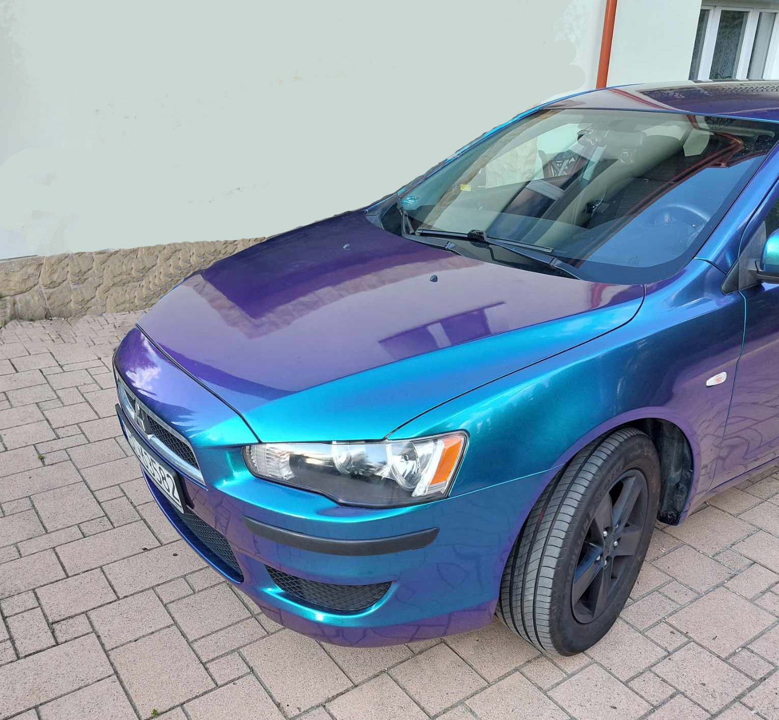 Mitsubishi Lancer z folią zmieniającą kolor od niebieskiego do fioletowego, zaparkowany na brukowanym podjeździe.
