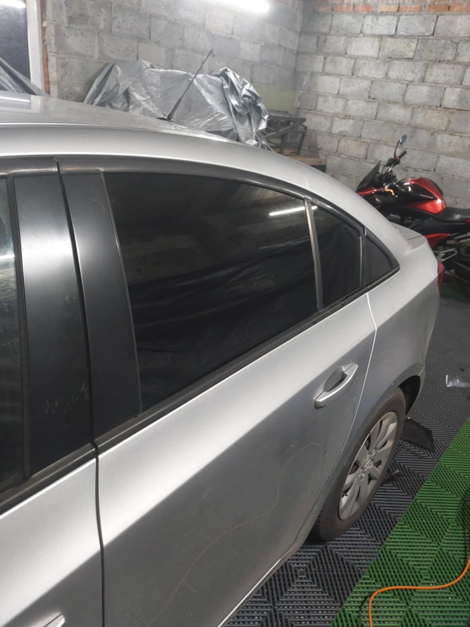 Srebrny samochód z przyciemnionymi szybami tylnymi, zaparkowany w warsztacie z widocznym motocyklem w tle.