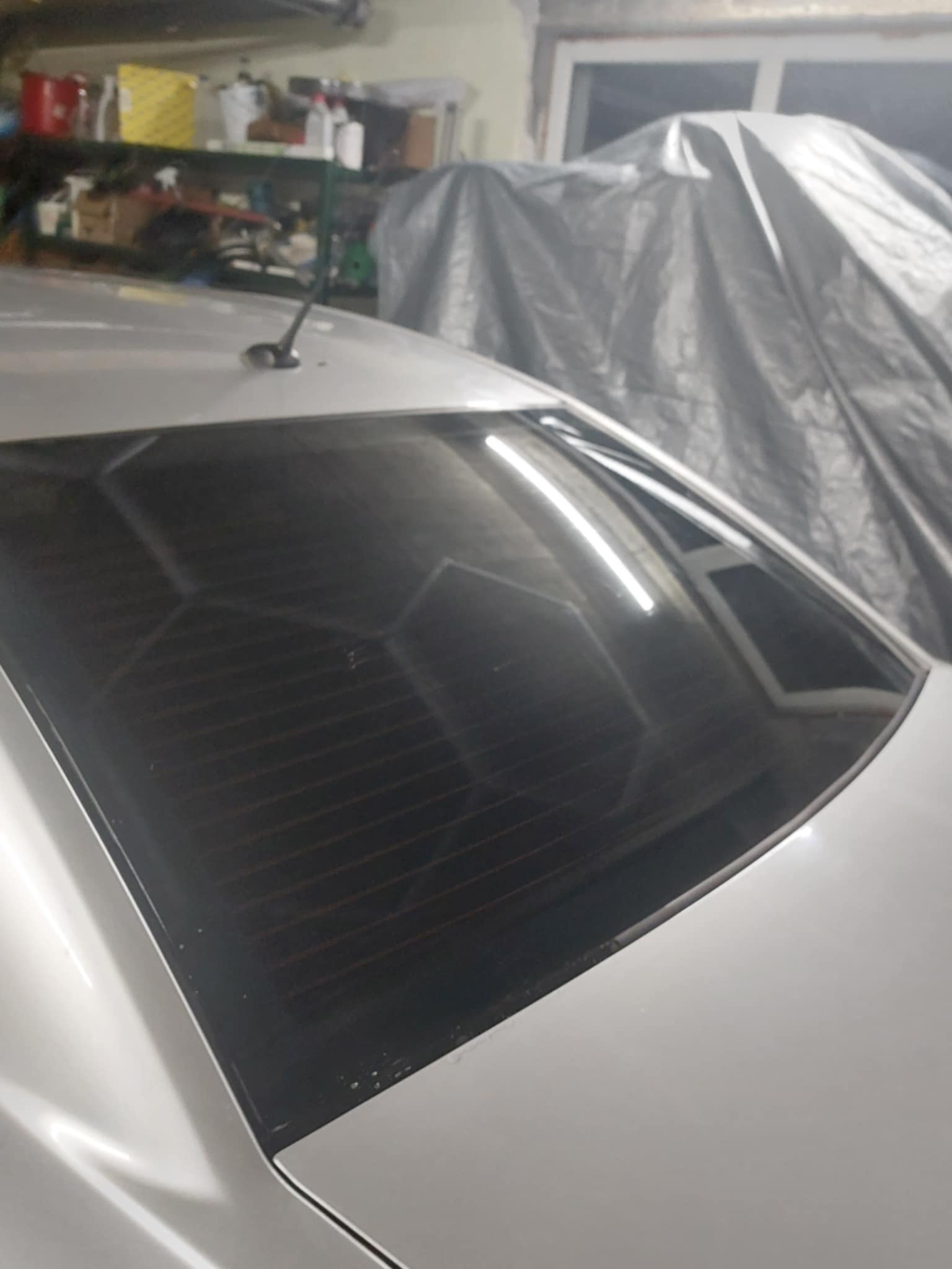 Przyciemniona tylna szyba białego samochodu, widoczne delikatne zarysowania na lakierze i tło garażowe z przykrytym plandeką obiektem.