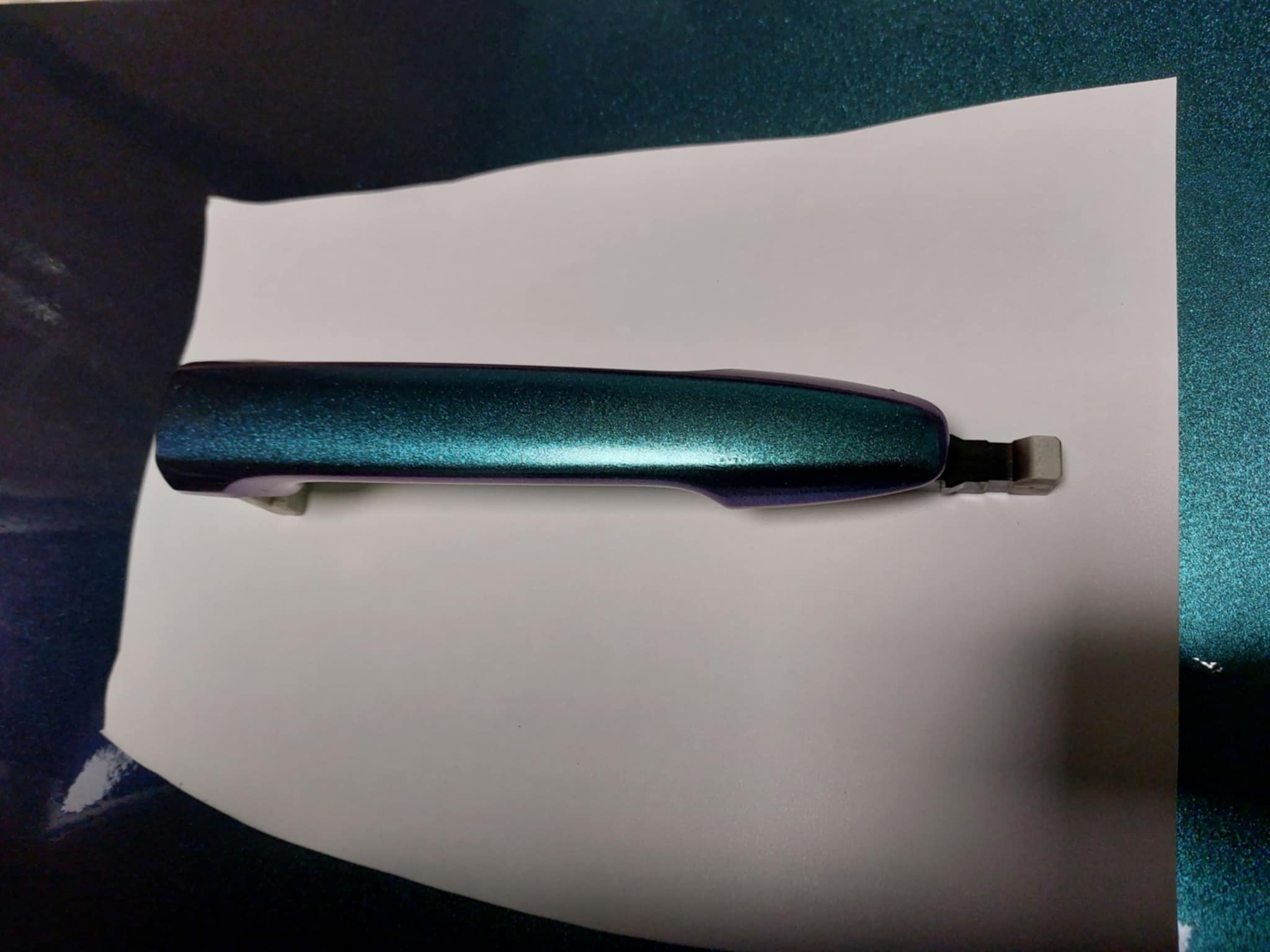 Pomalowana na kolor kameleona klamka samochodowa leży na białym arkuszu papieru, z boku widoczny fragment metalicznej powierzchni.