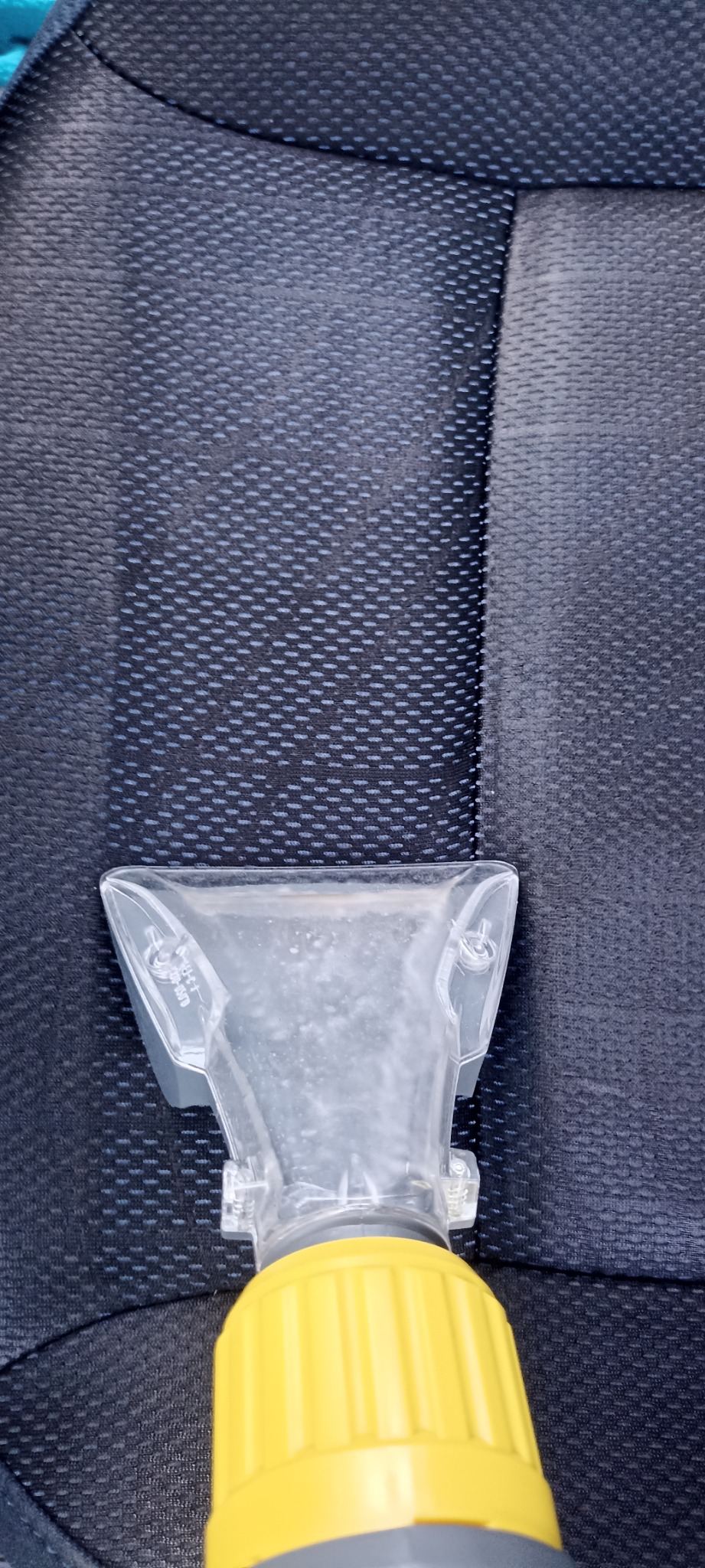 Dysza odkurzacza piorącego z żółtą rurą na ciemnej, teksturowanej tapicerce samochodowej, widoczna różnica w czystości między wypraną a niewypraną częścią.