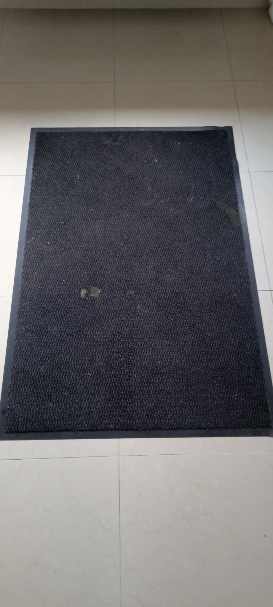 Ciemnoszara wycieraczka z gumową obwódką leżąca na jasnych, kwadratowych płytkach podłogowych, widoczne drobne zabrudzenia na powierzchni.