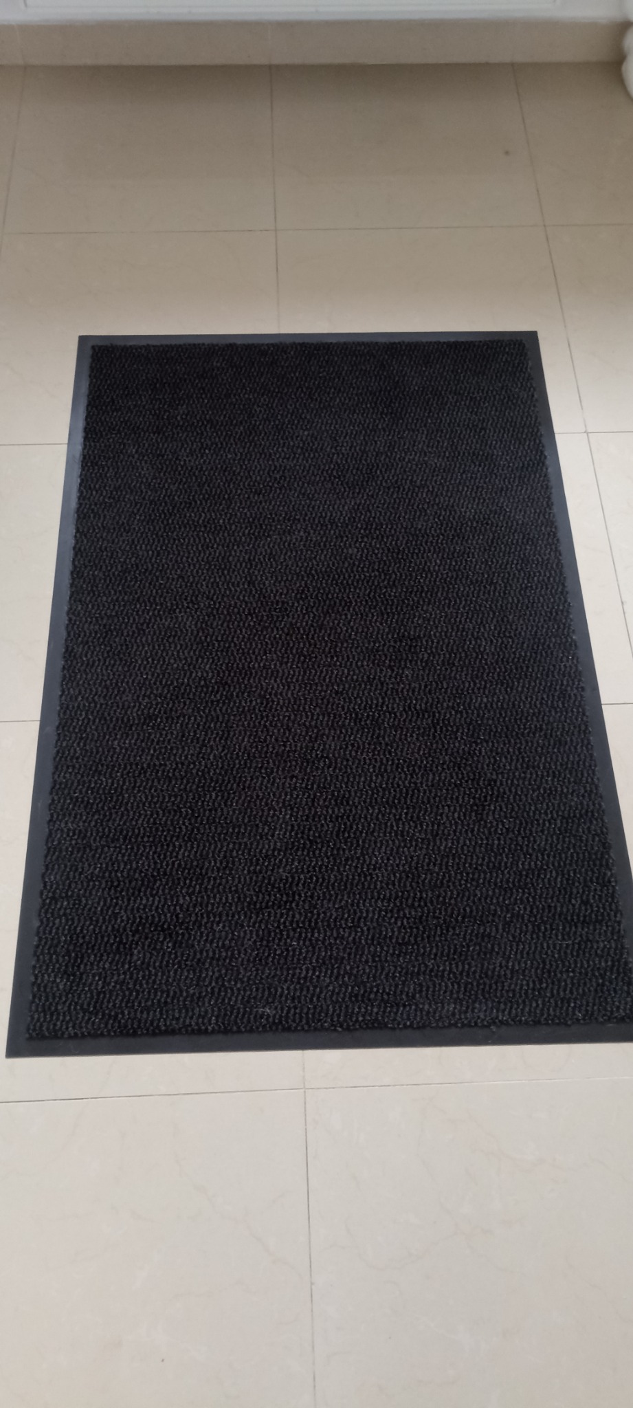 Ciemnoszary, prostokątny wycieraczka na beżowej podłodze z płytek, widok z góry.