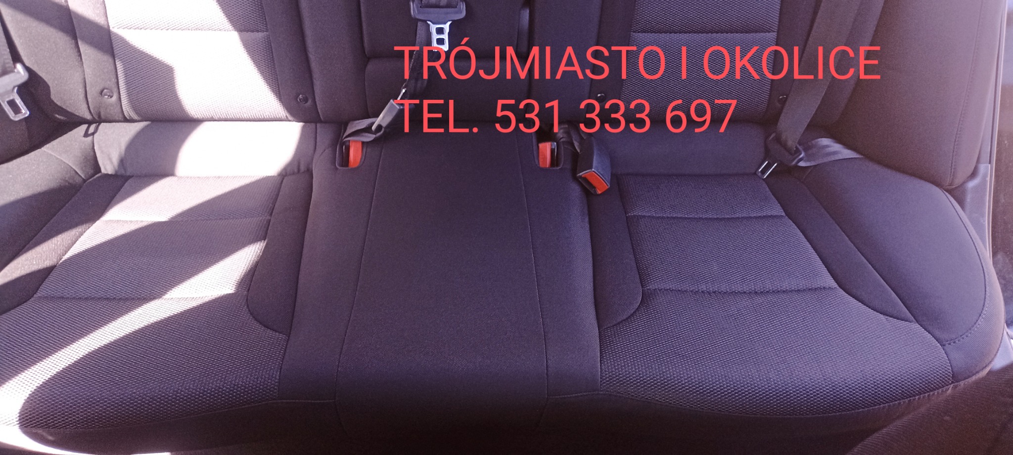 Ciemna, teksturowana tylna kanapa samochodowa z pasami bezpieczeństwa, z widocznym napisem 'TRÓJMIASTO I OKOLICE' oraz numerem telefonu.