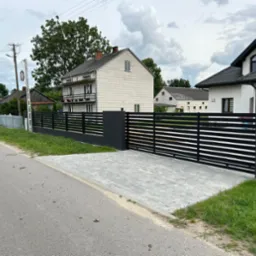 Nowoczesna, pozioma brama wjazdowa i przęsła ogrodzeniowe w kolorze antracytowym, z szarej kostki brukowej podjazd, w tle domy jednorodzinne.