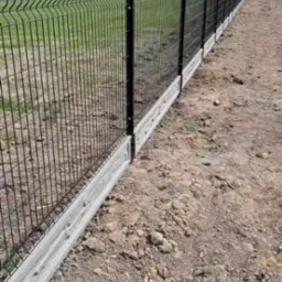 Długi, prosty odcinek czarnego ogrodzenia panelowego z podmurówką betonową, widok z boku, teren jeszcze niezagospodarowany.